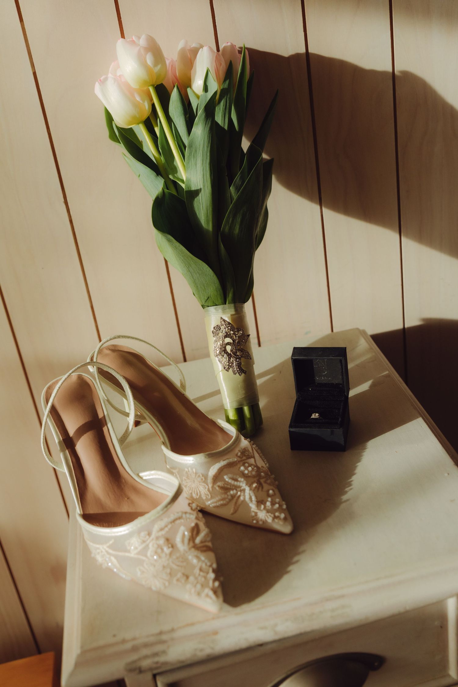 Bridal accessories flat lay featuring lace heels, pink tulip bouquet with brooch, and diamond ring box.