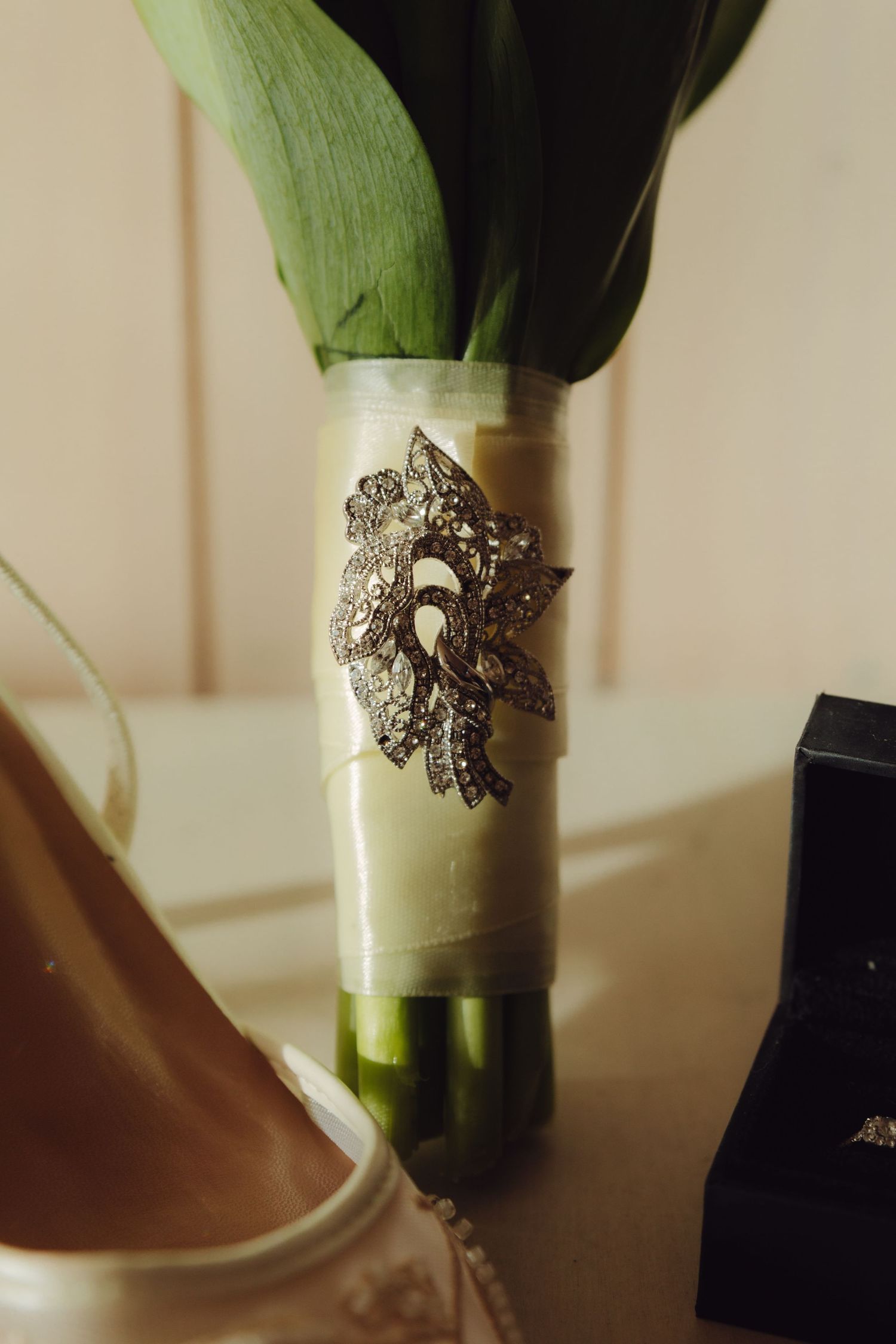 Elegant bridal bouquet wrapped in ribbon adorned with a crystal brooch beside wedding shoes and jewelry box.