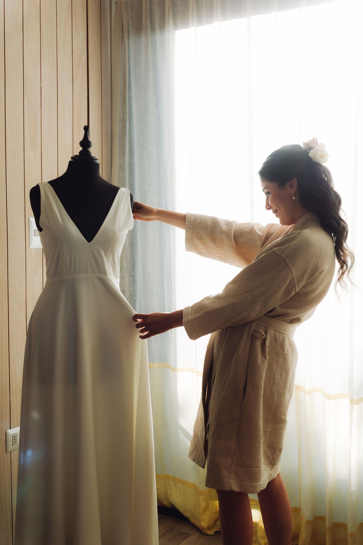 Brunette bride in linen robe lovingly adjusts her elegant white wedding dress on a mannequin by window.