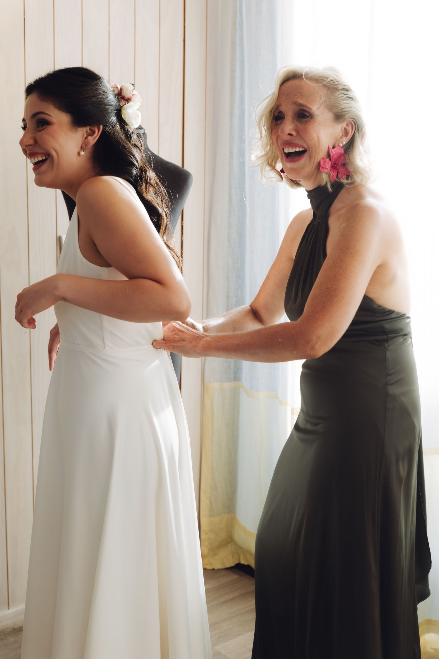 Joyful bride in white gown laughs as older woman in black dress fastens her wedding dress buttons.