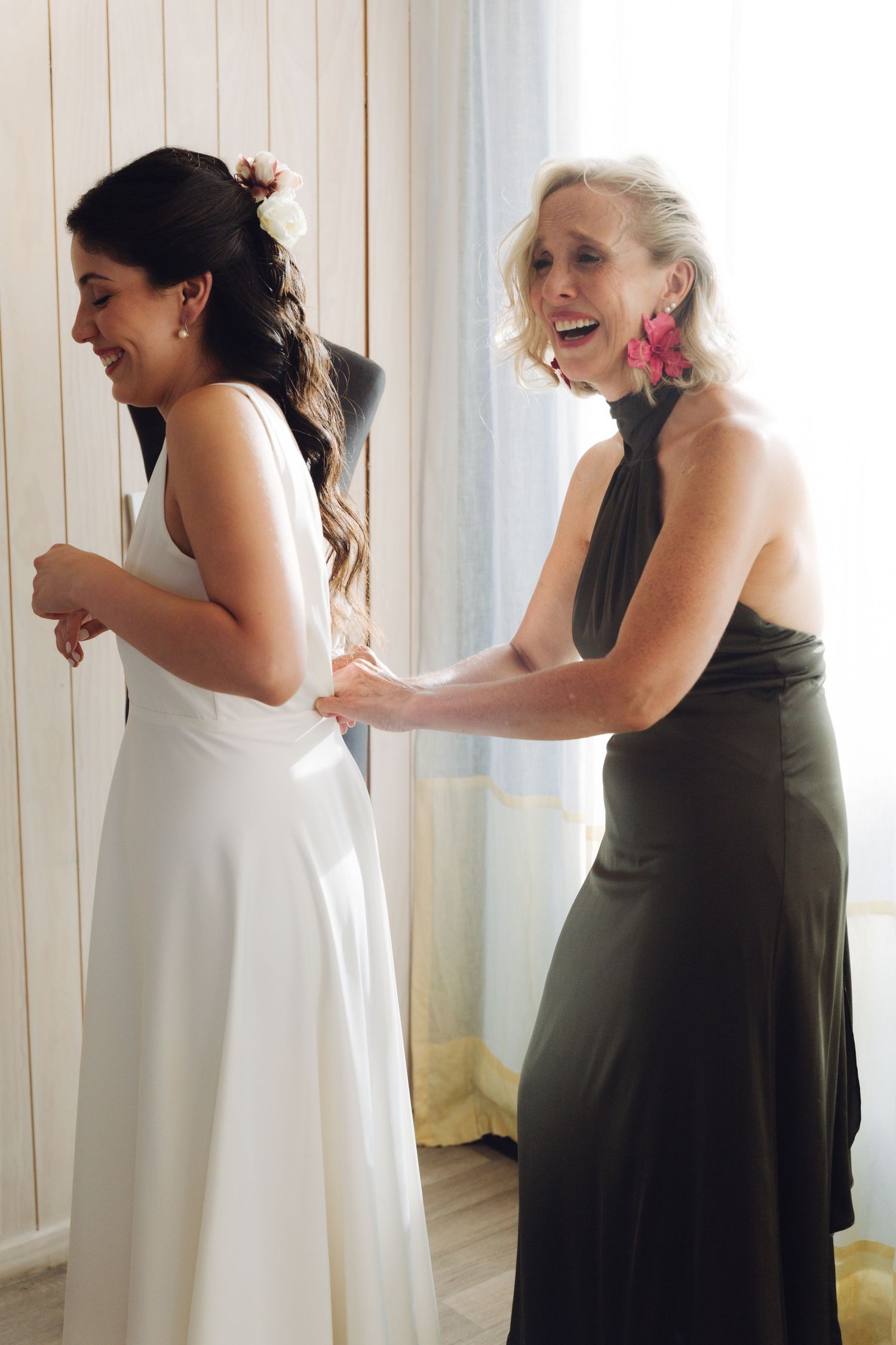 Smiling bride in white wedding gown gets dress fastened by laughing blonde woman in elegant black dress.