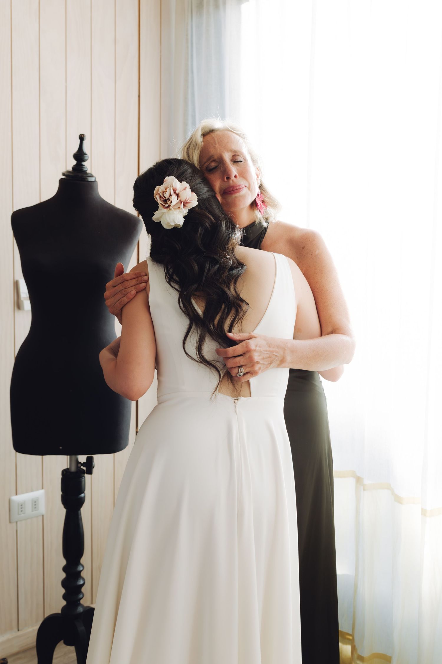 Emotional blonde woman in black dress tenderly embraces brunette bride in white gown near dress mannequin.