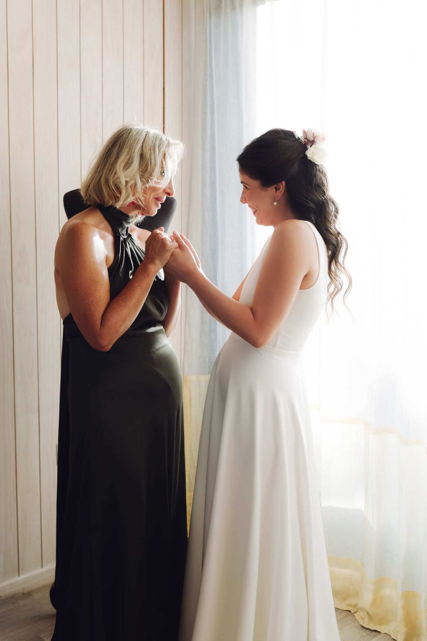 Bride in white halter gown holds hands with emotional blonde woman in black dress on wedding morning.
