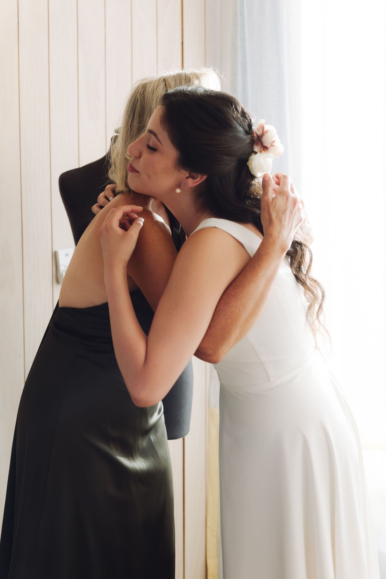Bride with floral hair accessory shares heartfelt hug with blonde woman in black dress before wedding ceremony.