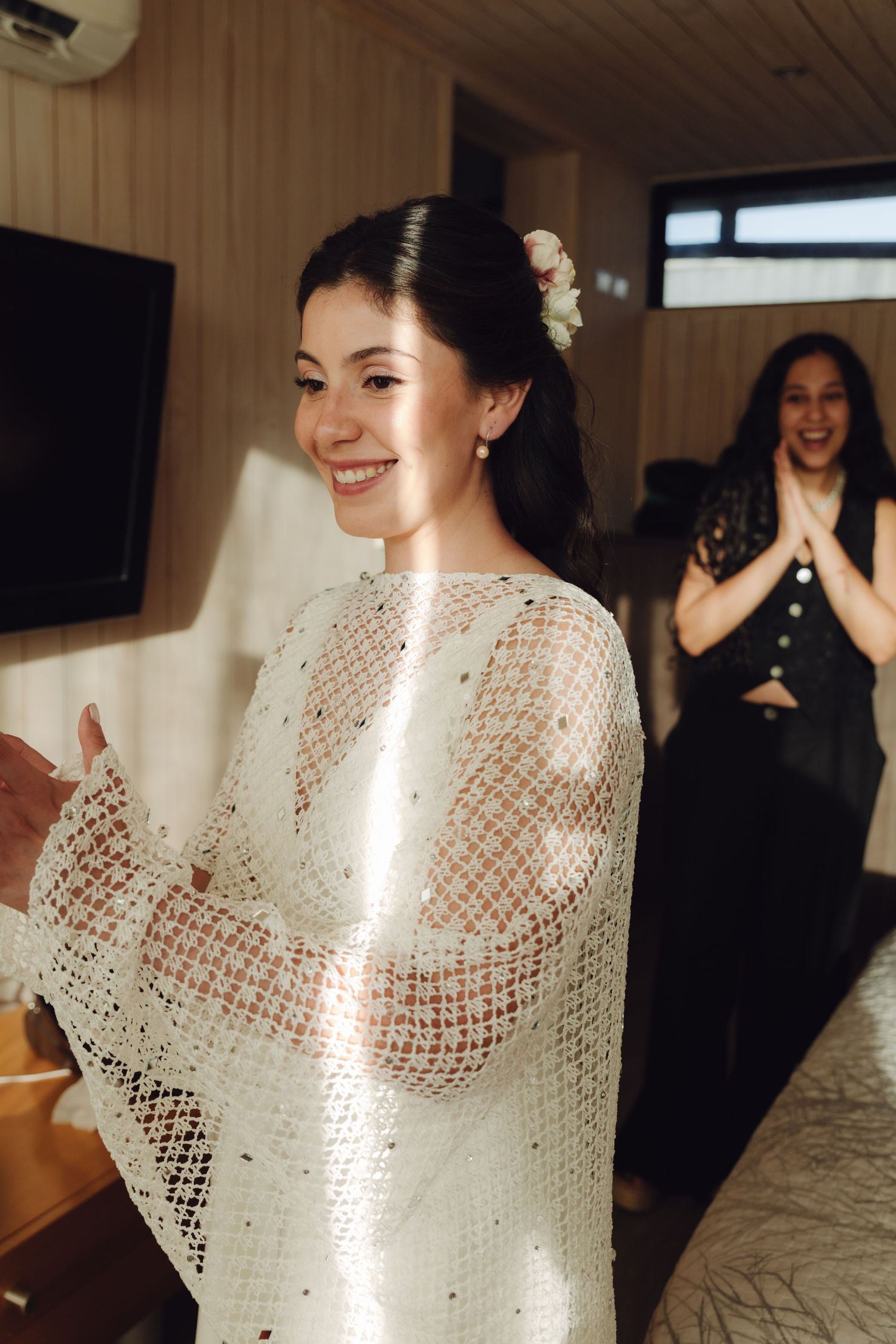A joyful bride in a white crochet cape smiling as her bridesmaid claps in delight.