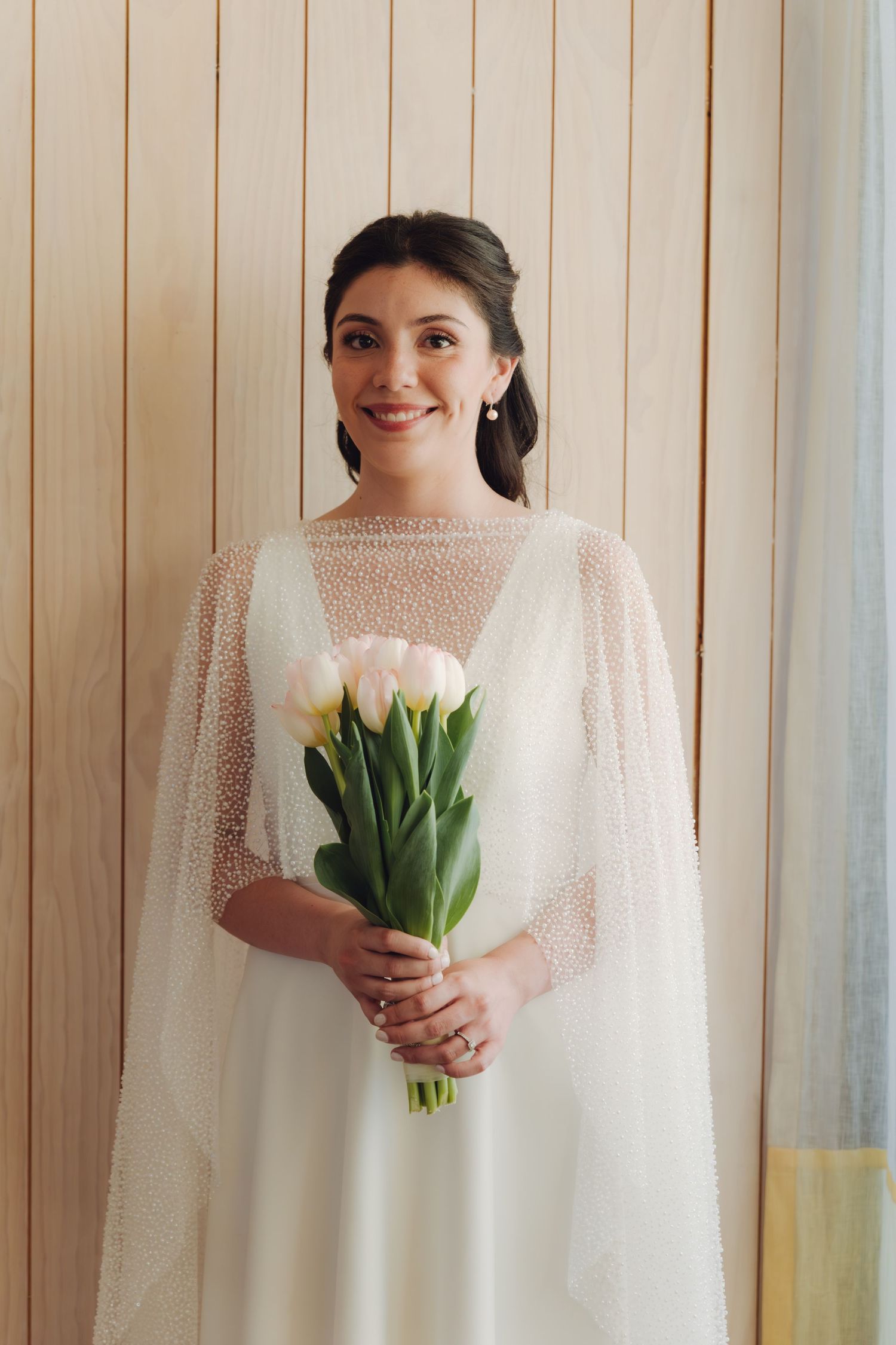 A smiling bride in a white beaded cape holding a bouquet of white tulips indoors.