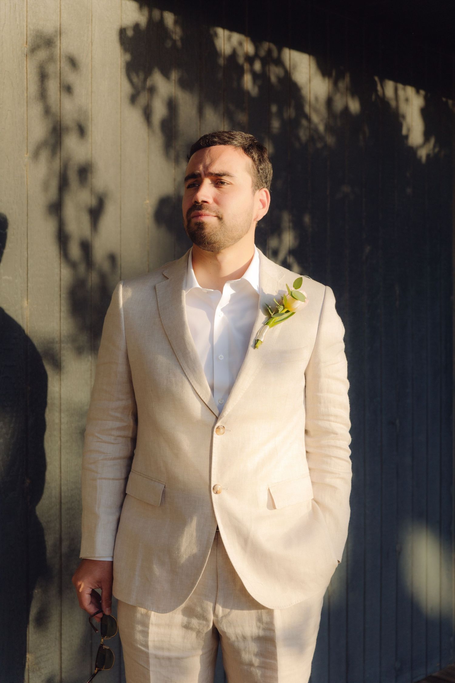 A handsome groom in a cream linen suit with a boutonniere standing in golden sunlight.
