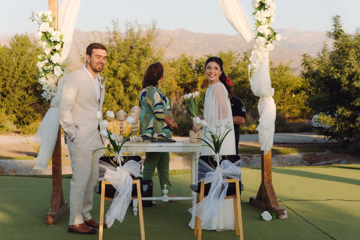 Bride and groom smiling at outdoor wedding ceremony altar with white draping and floral decorations.