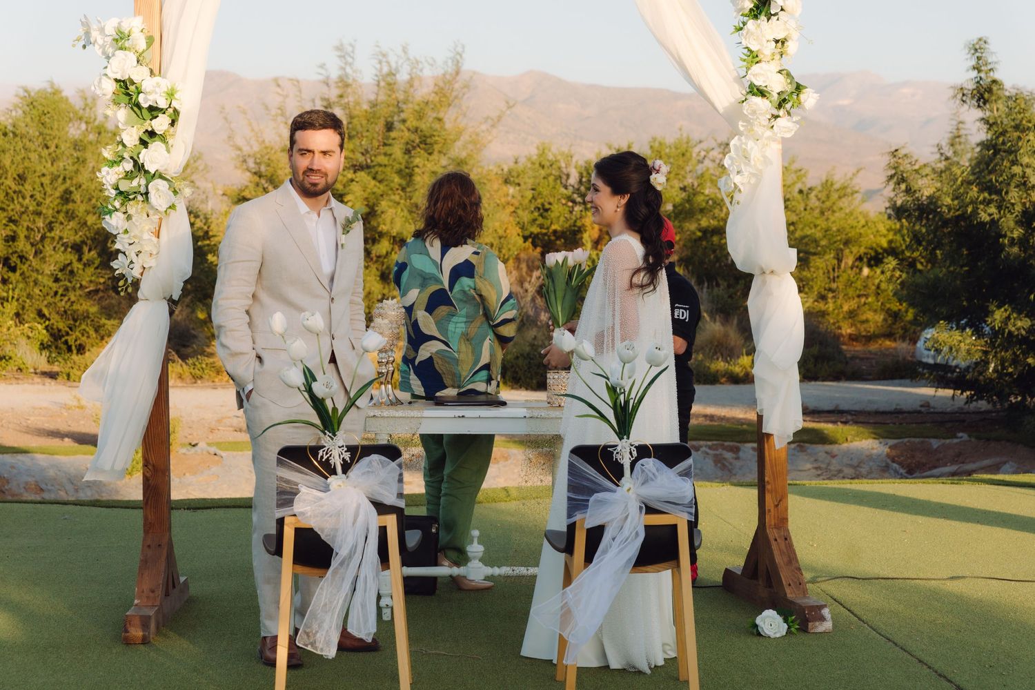 Couple exchanging vows at elegant outdoor wedding ceremony beneath wooden arch with white fabric.