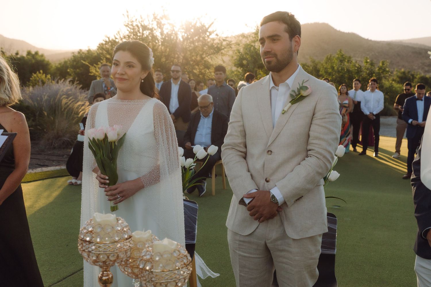 Bride holding white tulip bouquet stands beside groom in beige suit at golden hour wedding.