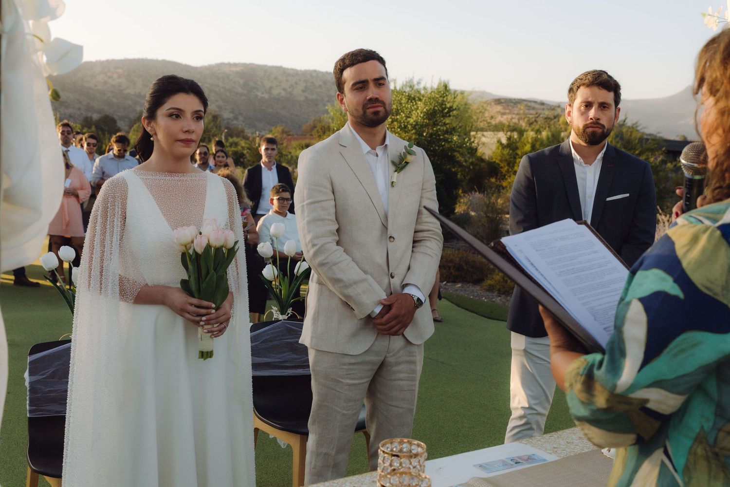 Bride with white tulips and groom in tan suit listen to officiant during outdoor wedding ceremony.