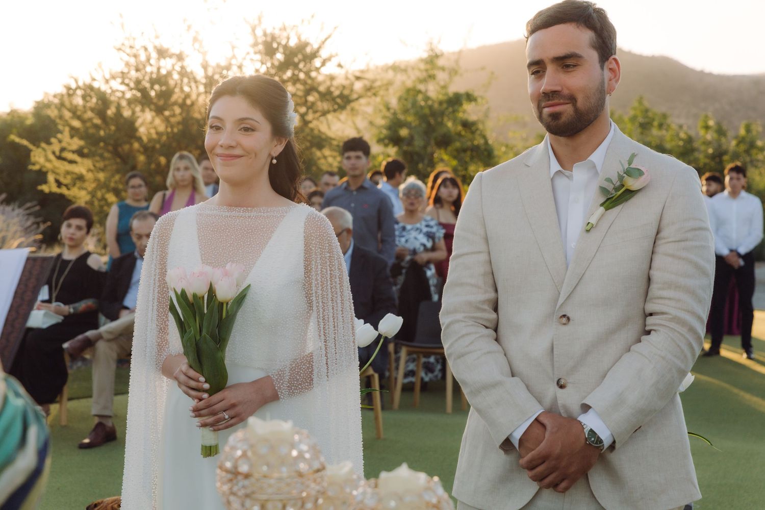 Smiling bride holding white tulips stands with groom at outdoor wedding ceremony with guests.