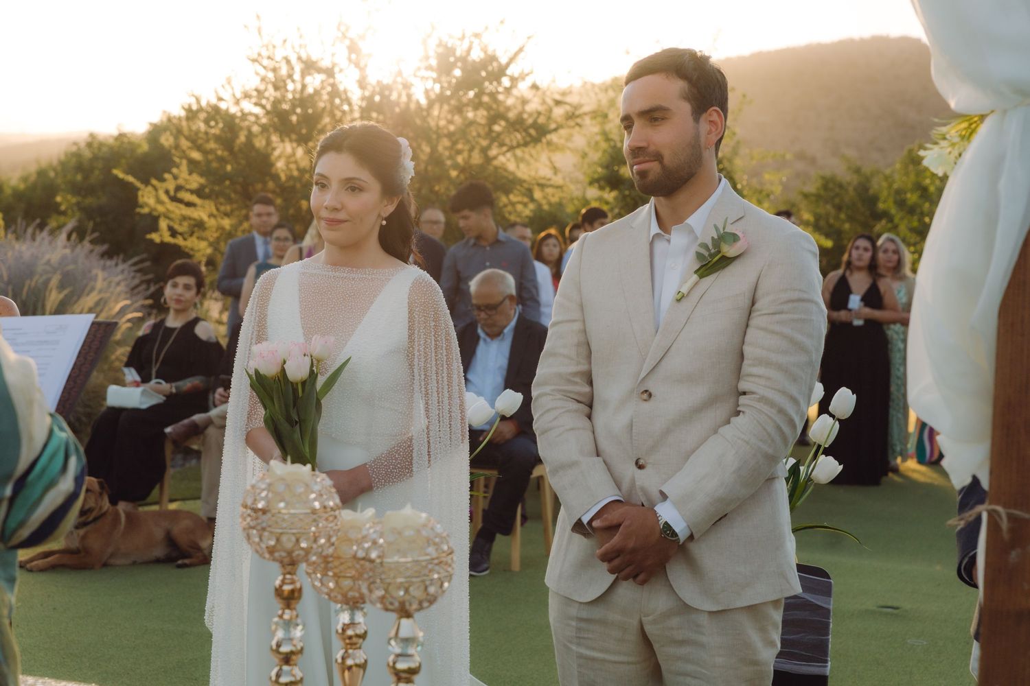 Bride carrying elegant bouquet with crystal accents stands beside groom at sunset wedding ceremony.