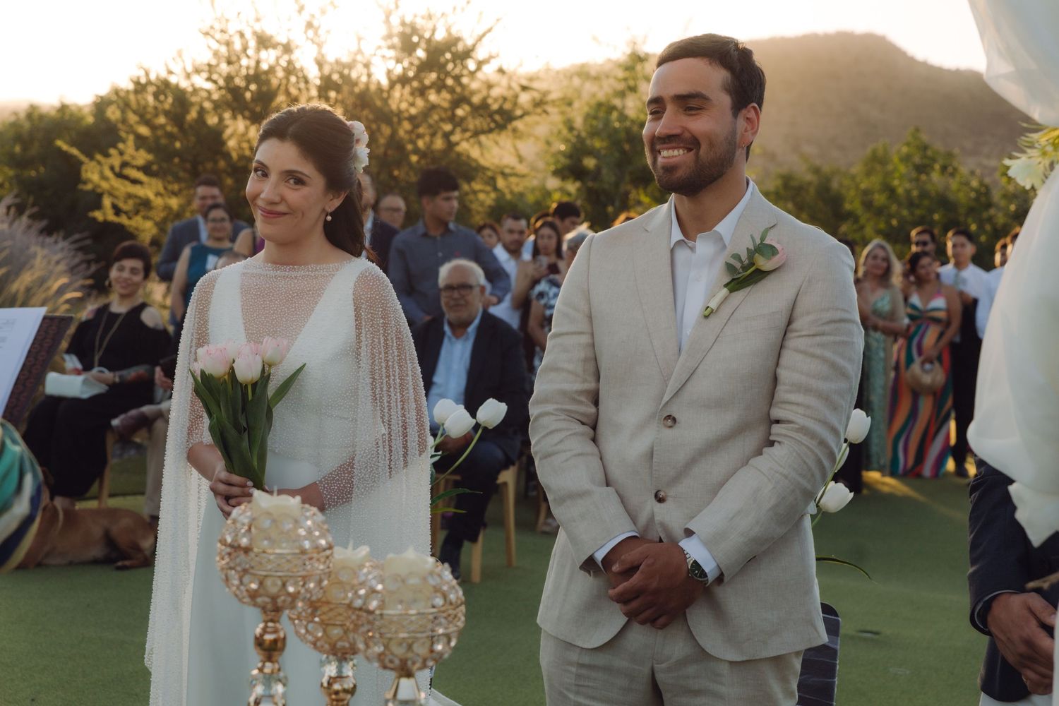 Joyful bride and groom smile during outdoor wedding ceremony surrounded by seated guests at dusk.