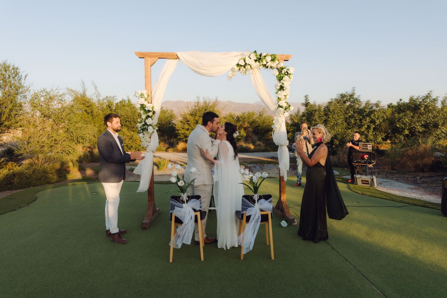 Newlyweds share first kiss under wooden floral arch at intimate outdoor wedding ceremony.