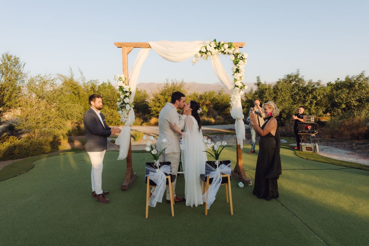 Couple kisses under white draped wedding arch on green lawn with small group of witnesses.