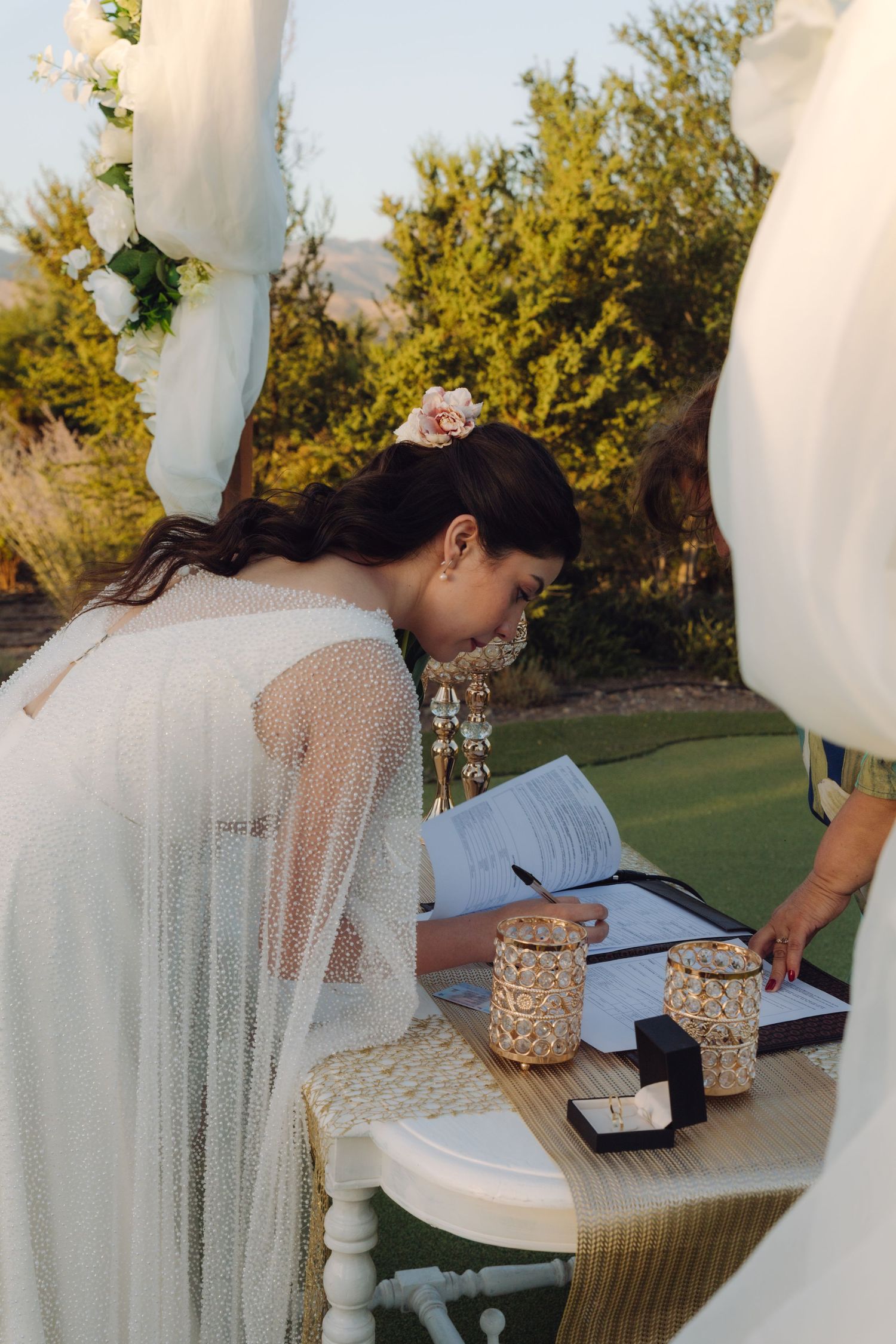 Bride in white lace gown signs marriage certificate at decorated table during outdoor ceremony.