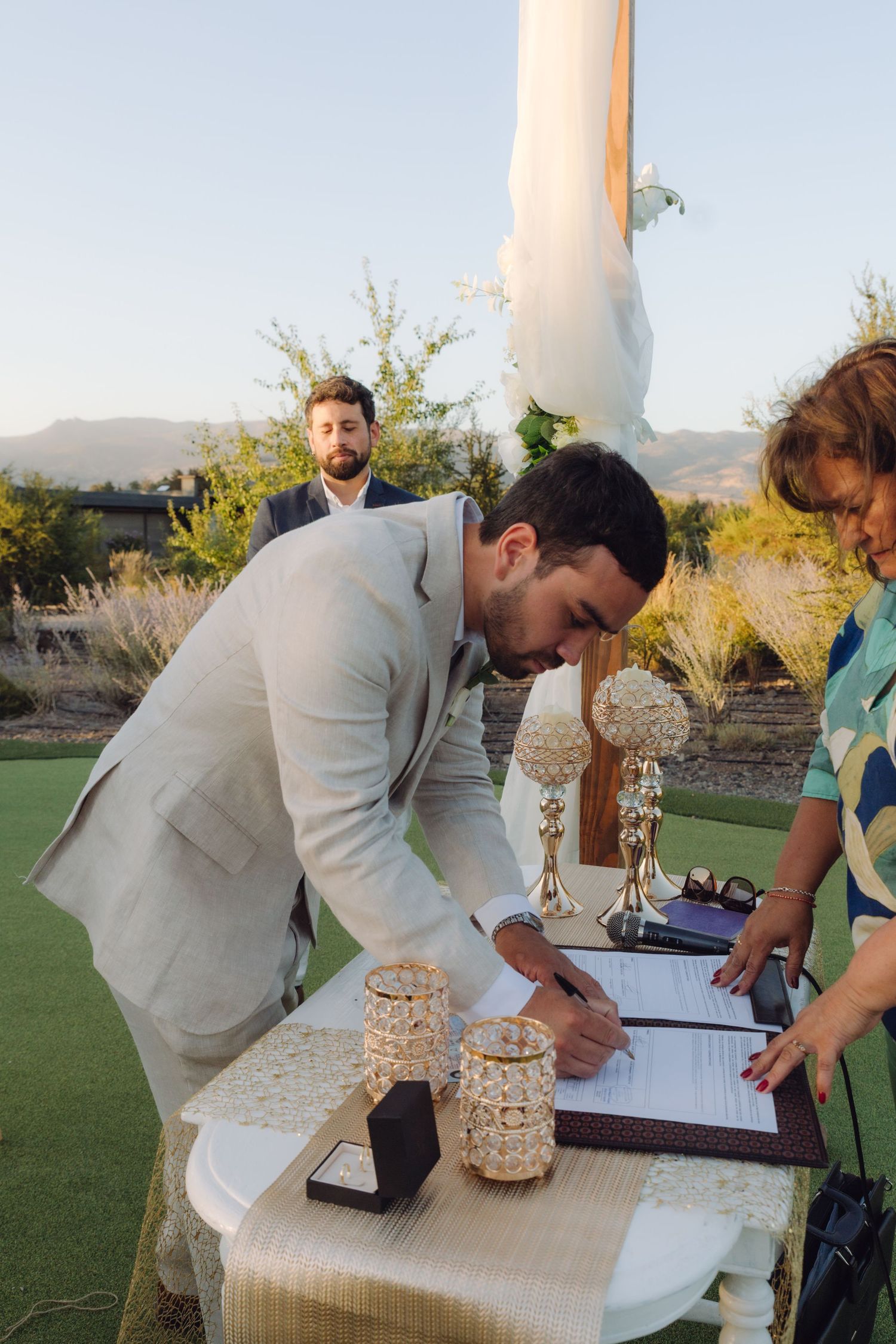 Groom in beige suit signing wedding documents at an outdoor ceremony table with candles.