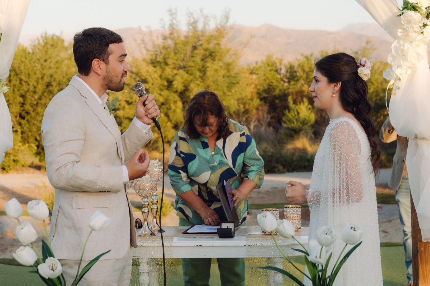 Groom speaks vows into microphone while bride smiles during outdoor wedding ceremony.