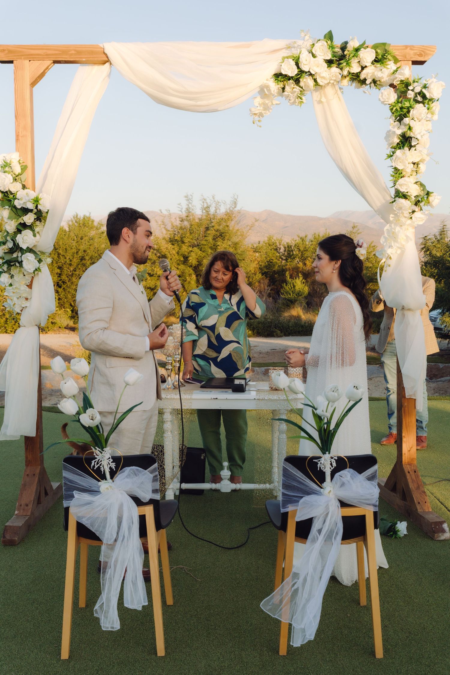 Couple exchanges vows under floral arch with white draping at outdoor wedding ceremony.