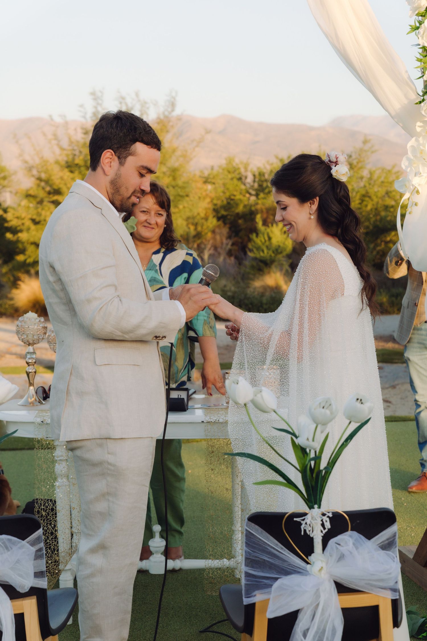 Groom places wedding ring on brides finger as officiant watches during outdoor ceremony.