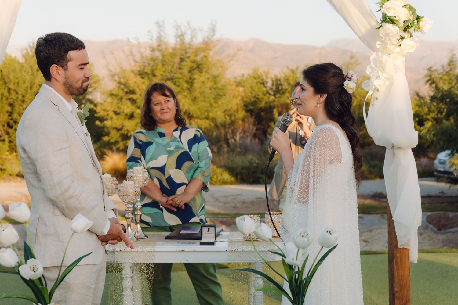 Bride reads vows while groom listens during outdoor wedding ceremony at golden hour.