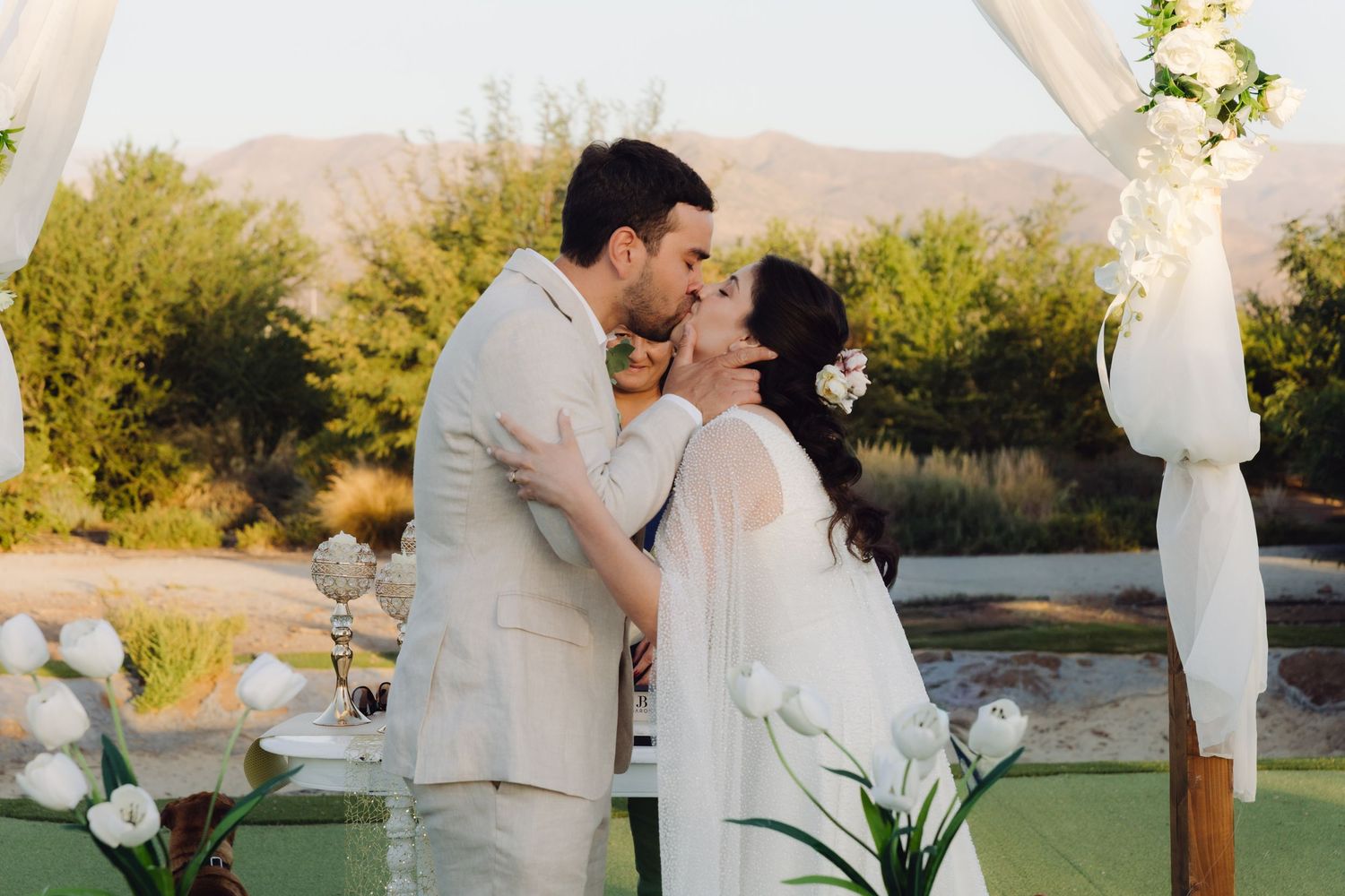 Newlyweds share first kiss under white draped arch with mountain backdrop at sunset.
