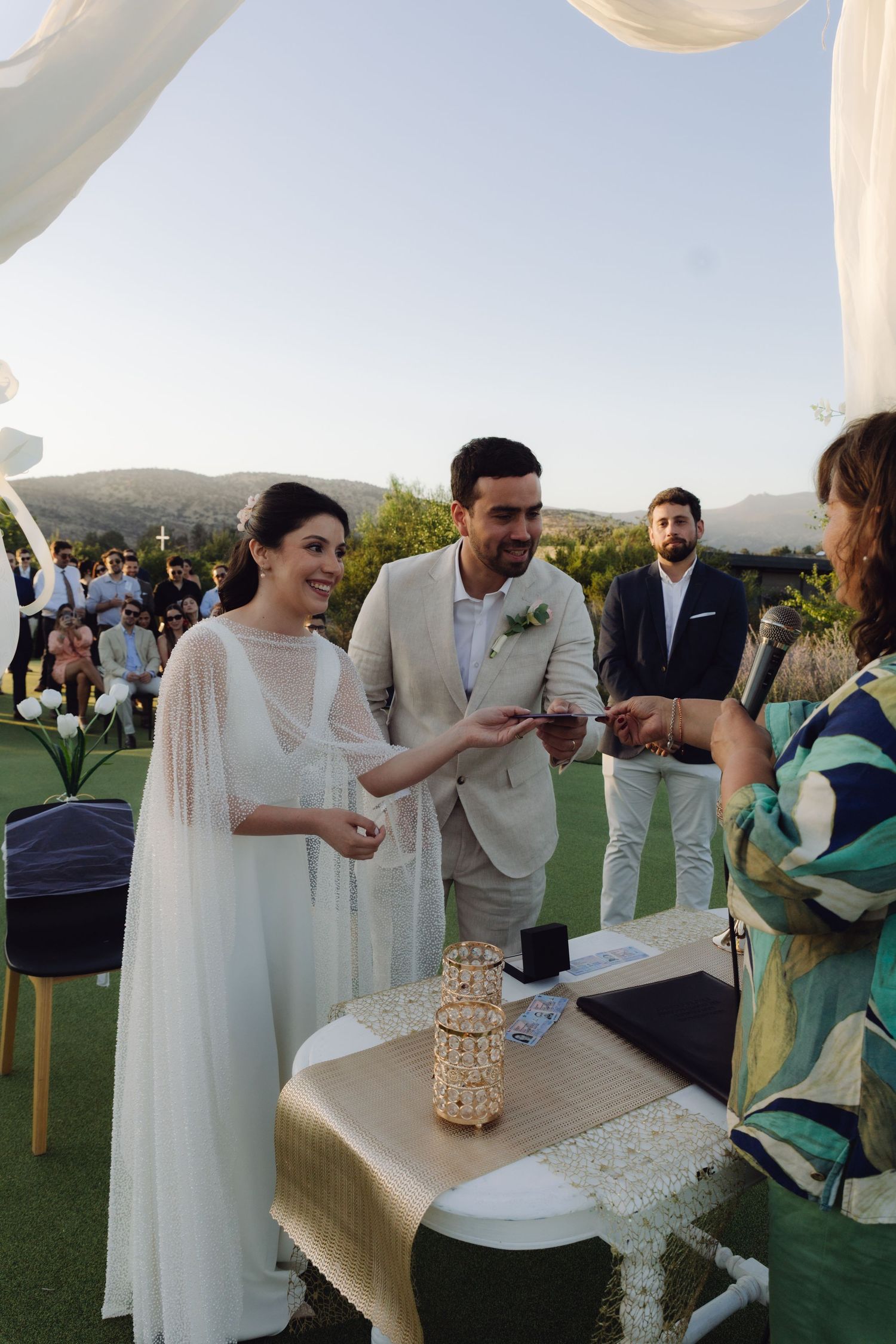 Bride and groom receive wedding rings from officiant during outdoor ceremony at dusk.