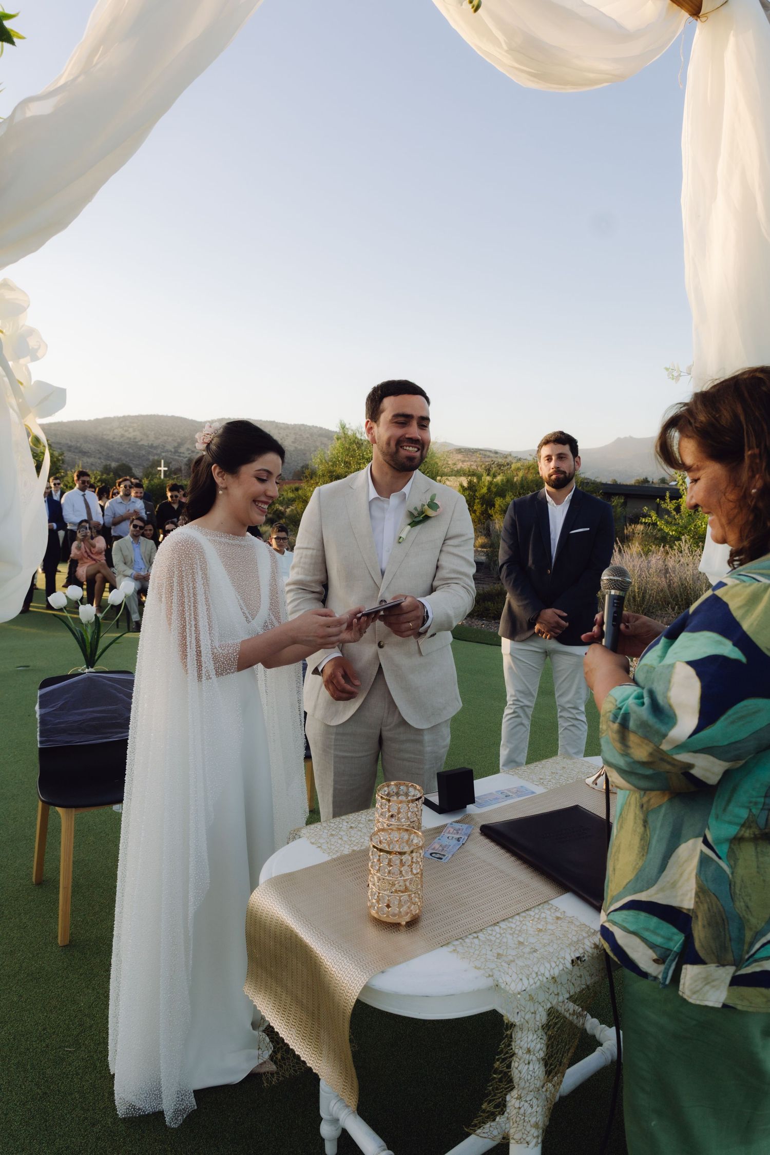 Smiling couple exchanges rings during outdoor wedding ceremony with mountain views behind them.