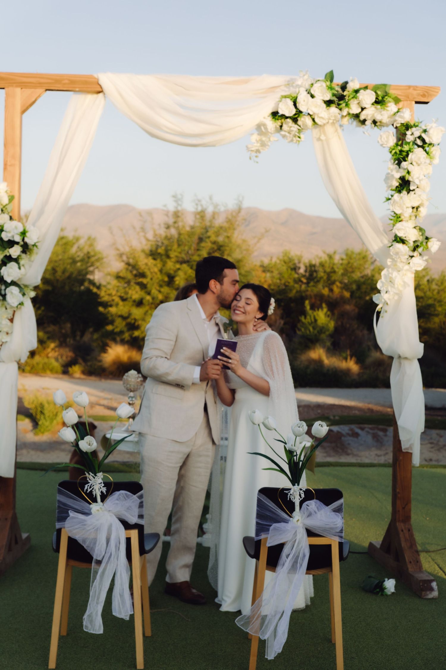 Newlyweds kiss and hold marriage certificate under floral arch after outdoor wedding ceremony.