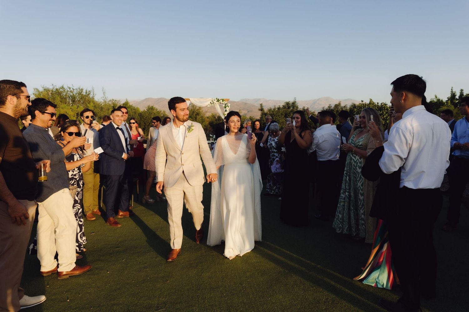 Bride and groom walk through cheering wedding guests outdoors with mountain views at golden hour.