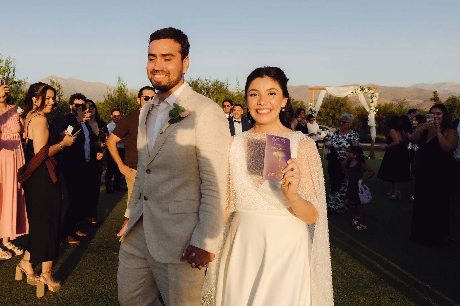 Smiling bride holds wedding booklets while standing with groom after outdoor ceremony at sunset.