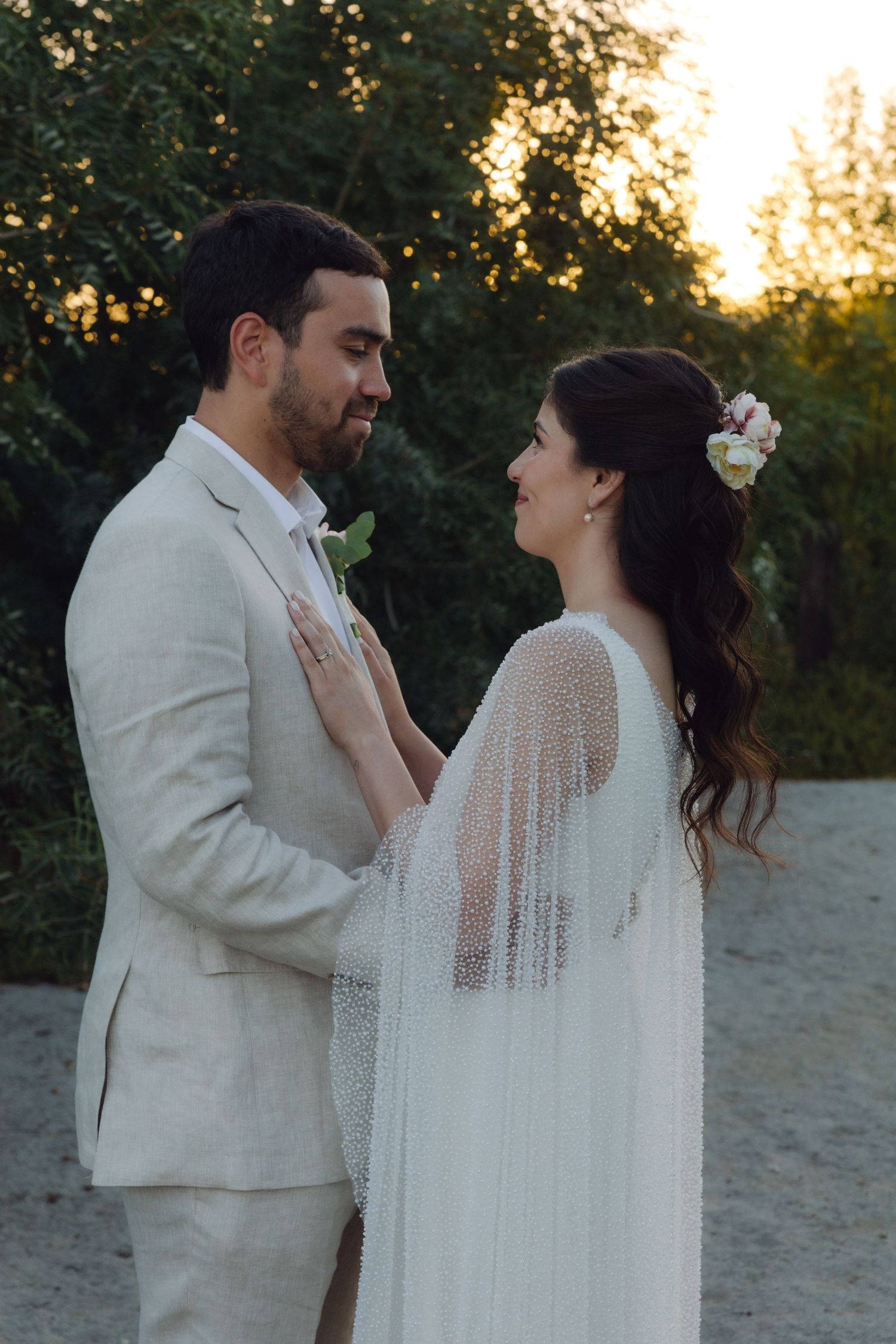 Bride in pearl cape wedding dress and groom share romantic moment at golden hour outdoors.