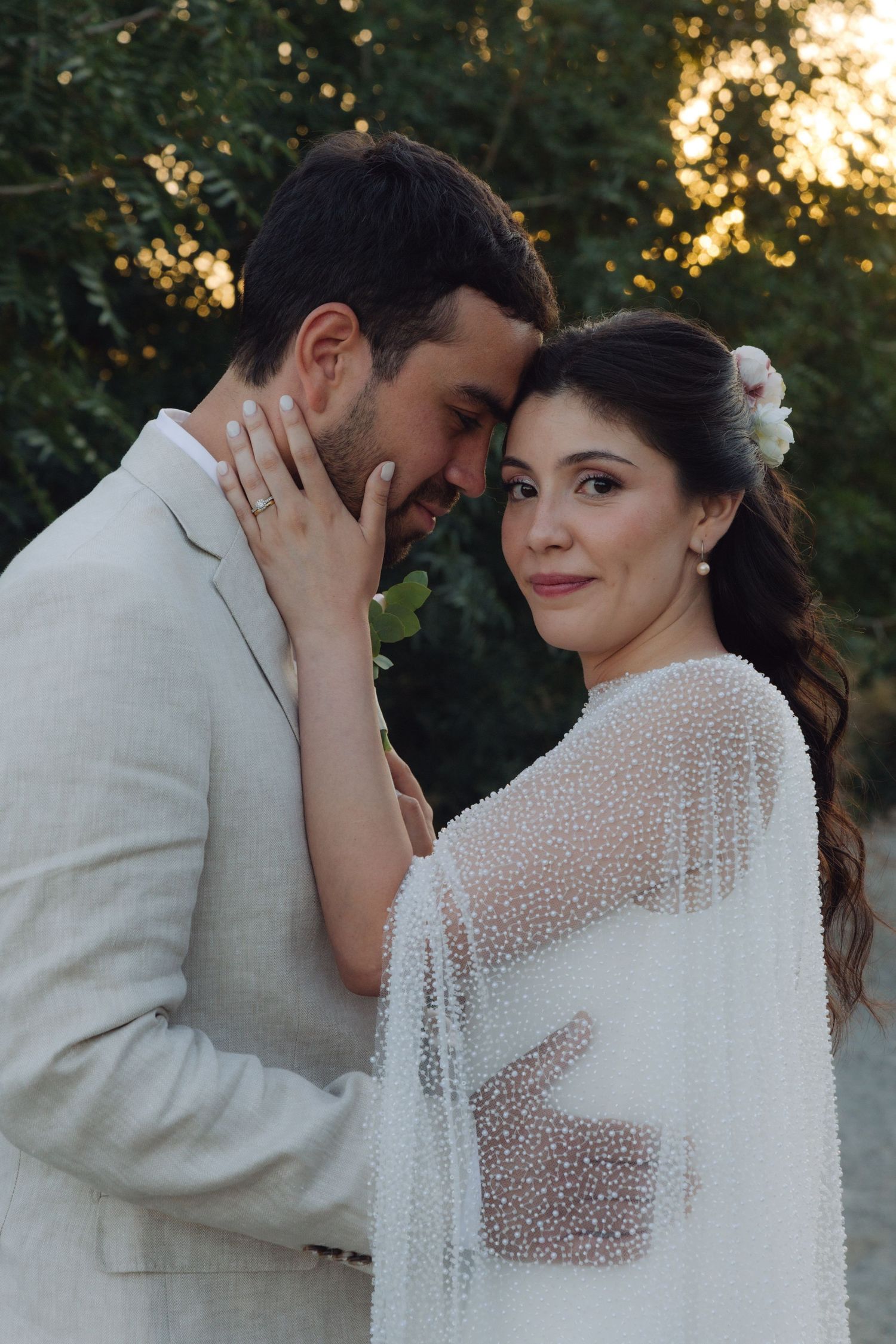Bride in beaded cape gown with floral hair accessory embraces groom at sunset wedding.