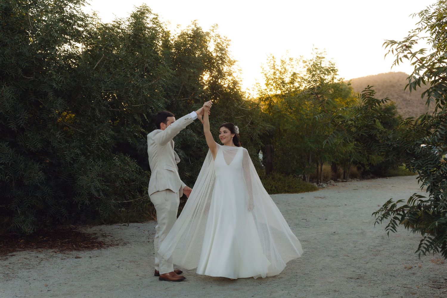 Groom twirls bride in flowing white cape wedding dress on sandy path at sunset.