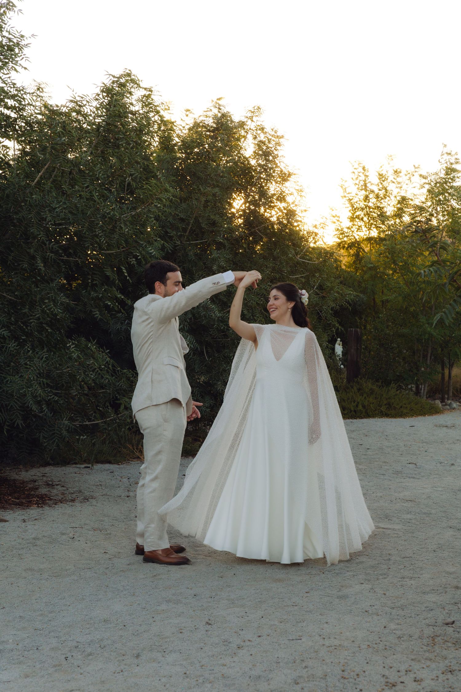 Groom spins bride in elegant white cape gown on sandy path surrounded by lush trees.