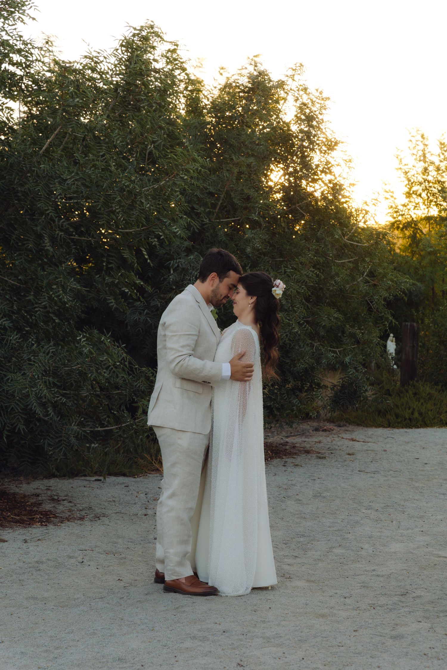 Bride and groom share a romantic embrace on sandy path surrounded by trees at sunset.