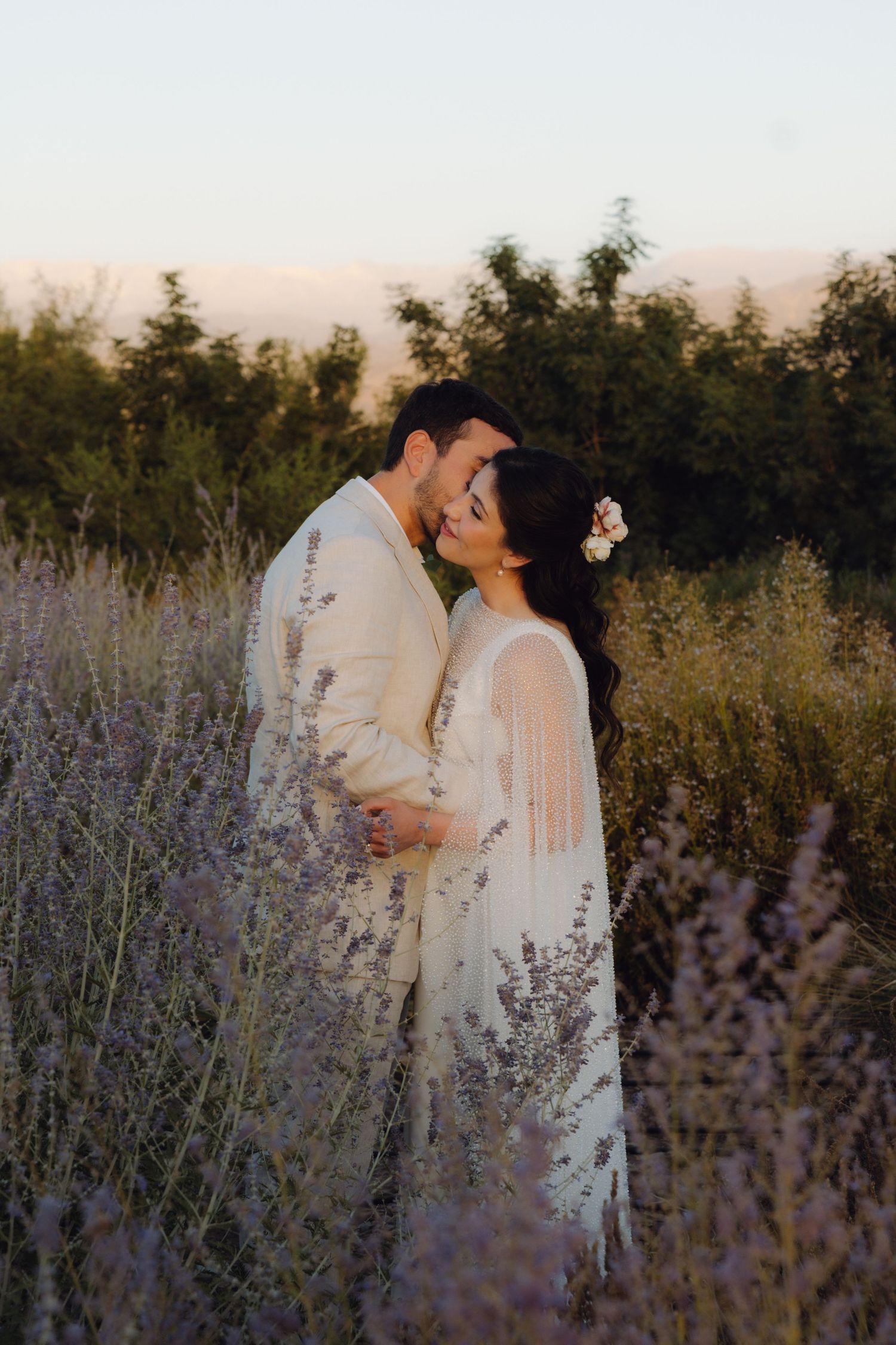 Bride and groom embrace romantically among lavender fields with mountain backdrop at golden hour.