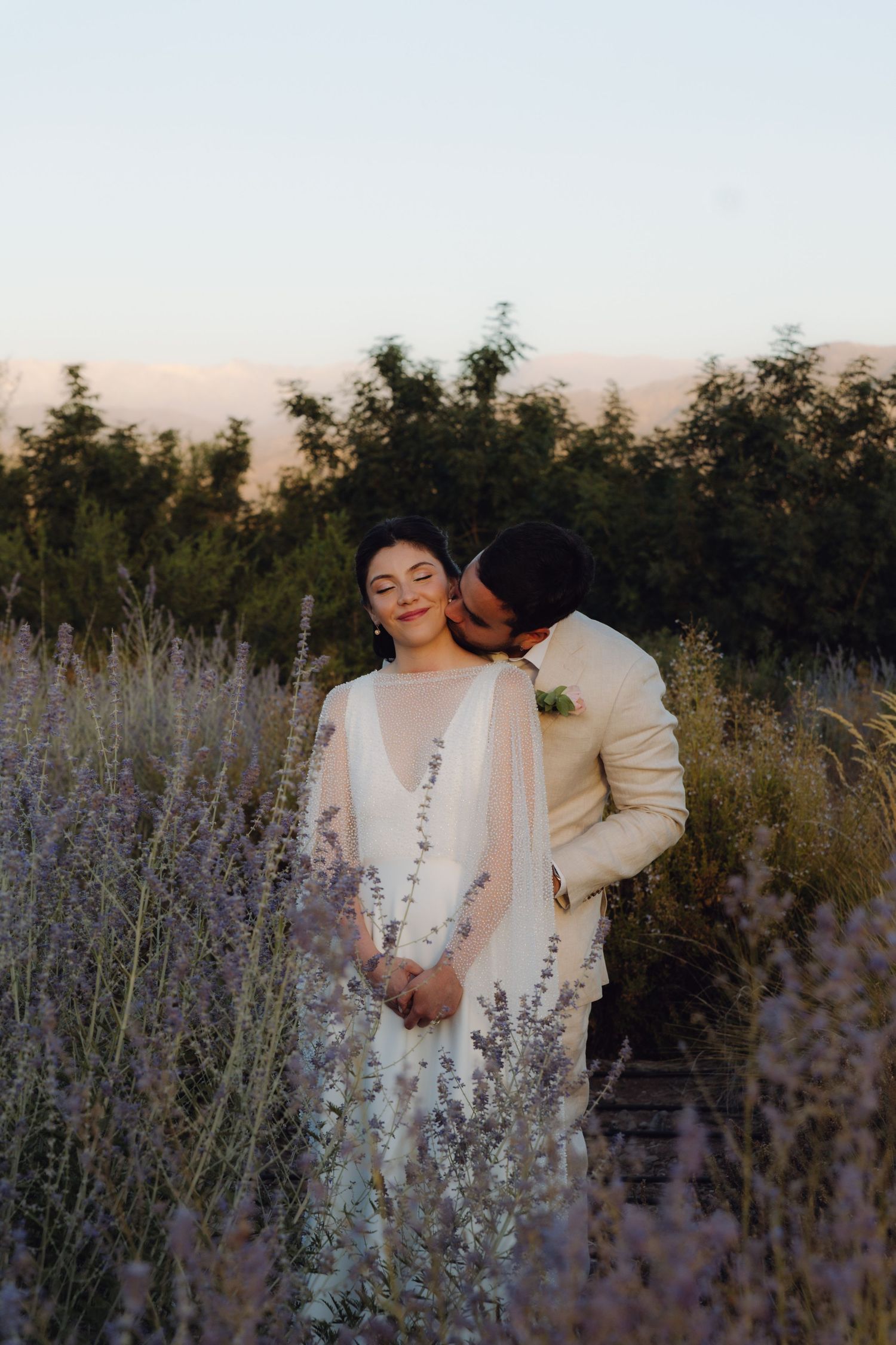 Groom kissing bride in lavender field at sunset, bride in white gown with sheer cape smiling blissfully.