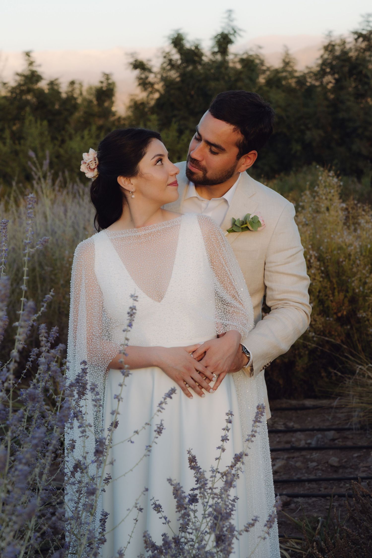 Wedding couple embracing in lavender field at dusk, bride in beaded cape gown gazing up at groom.