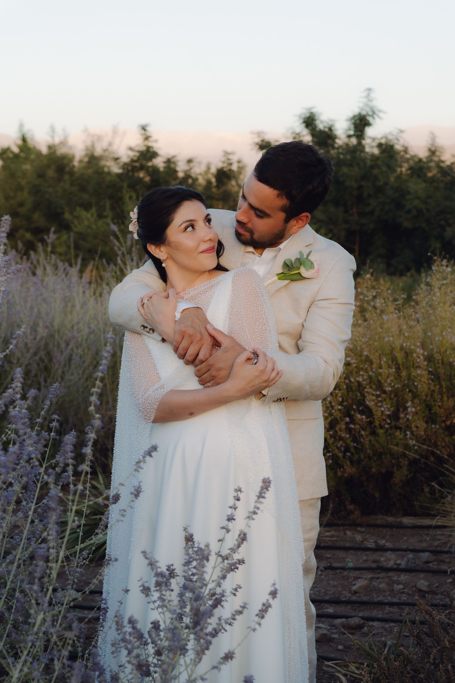 Bride and groom sharing tender embrace on wooden path through lavender field at golden hour.