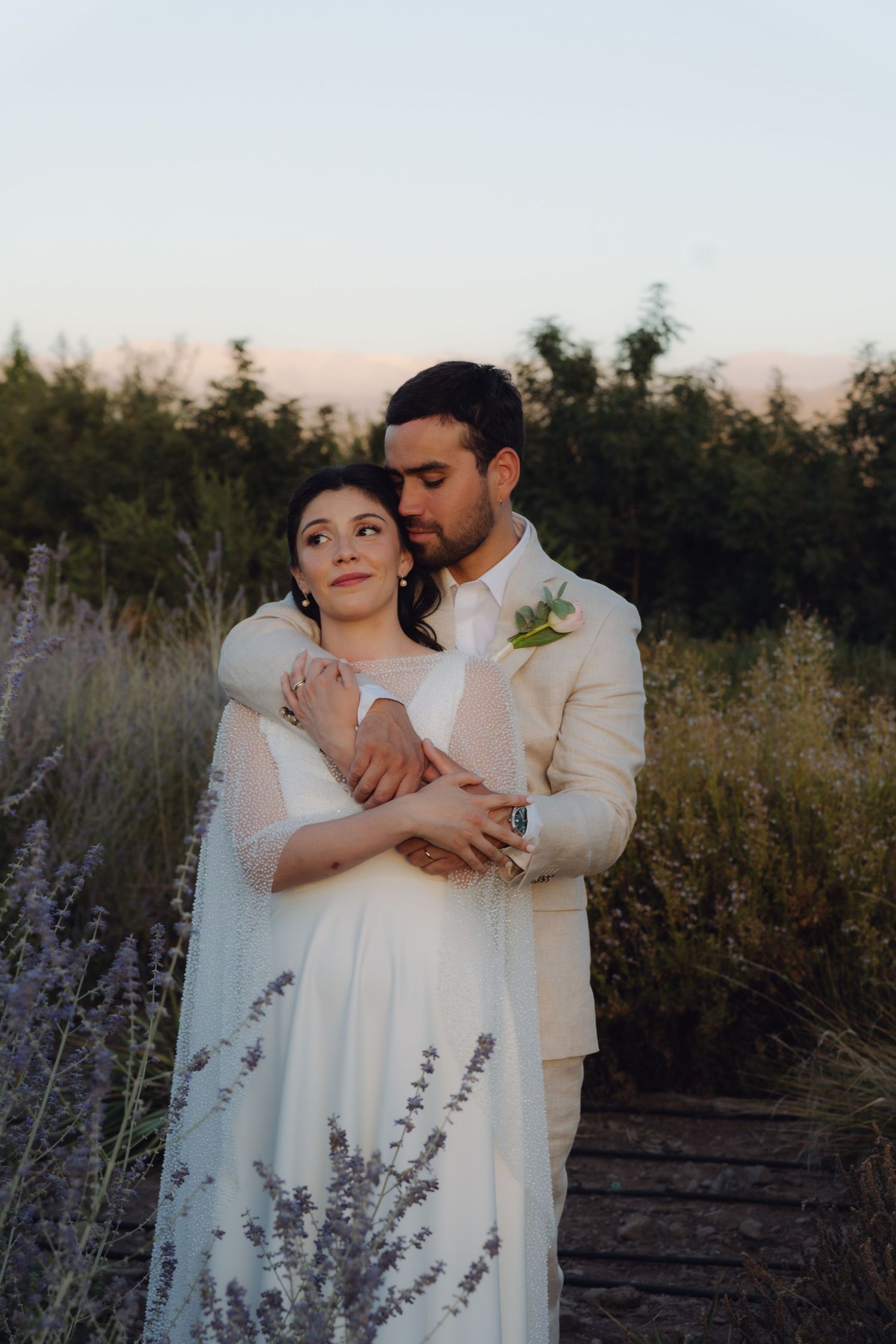 Groom tenderly embracing bride from behind in lavender field at sunset, both in elegant wedding attire.