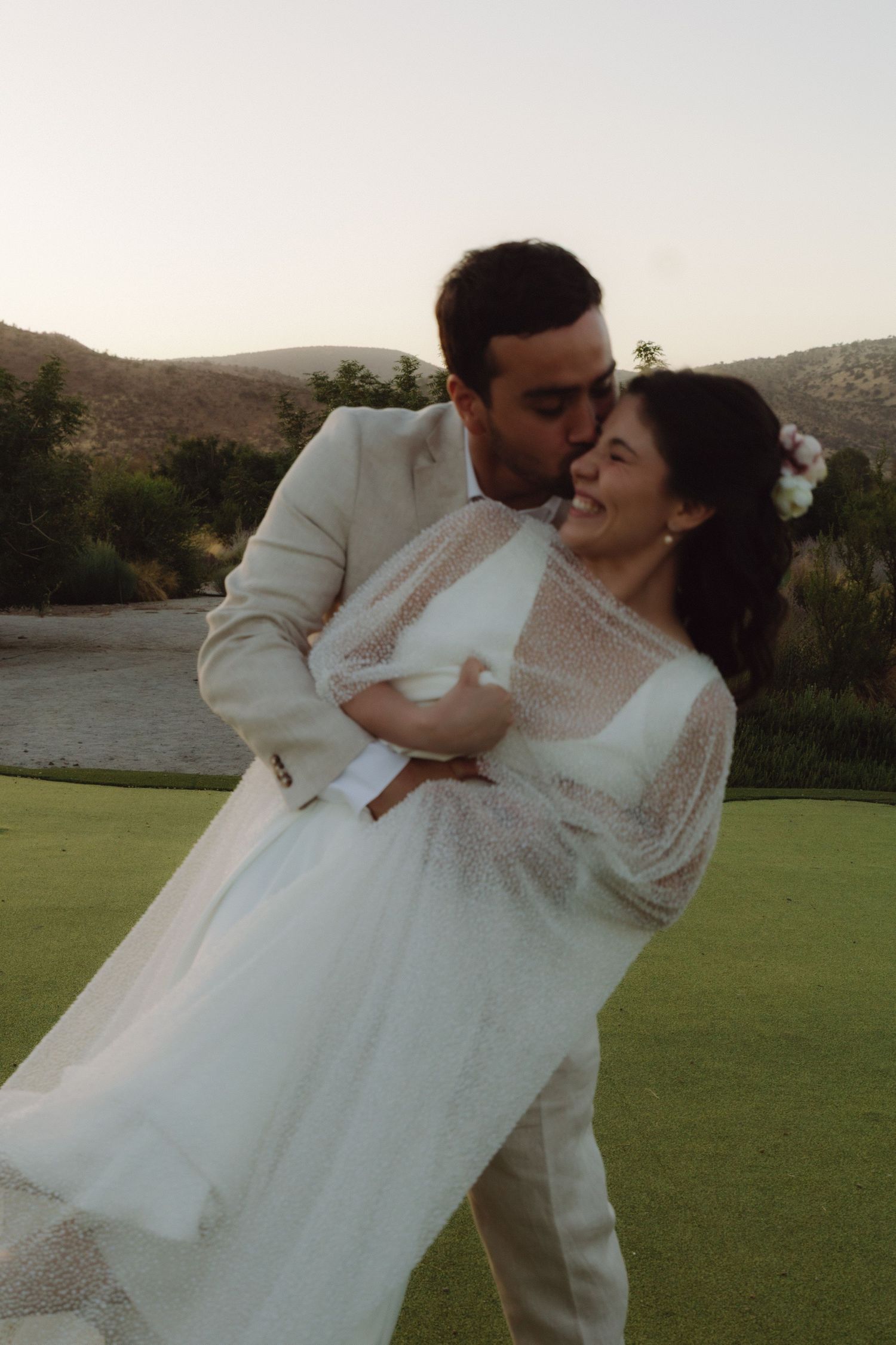 Groom dipping laughing bride on golf course at sunset, bride in flowing white gown with sheer cape.