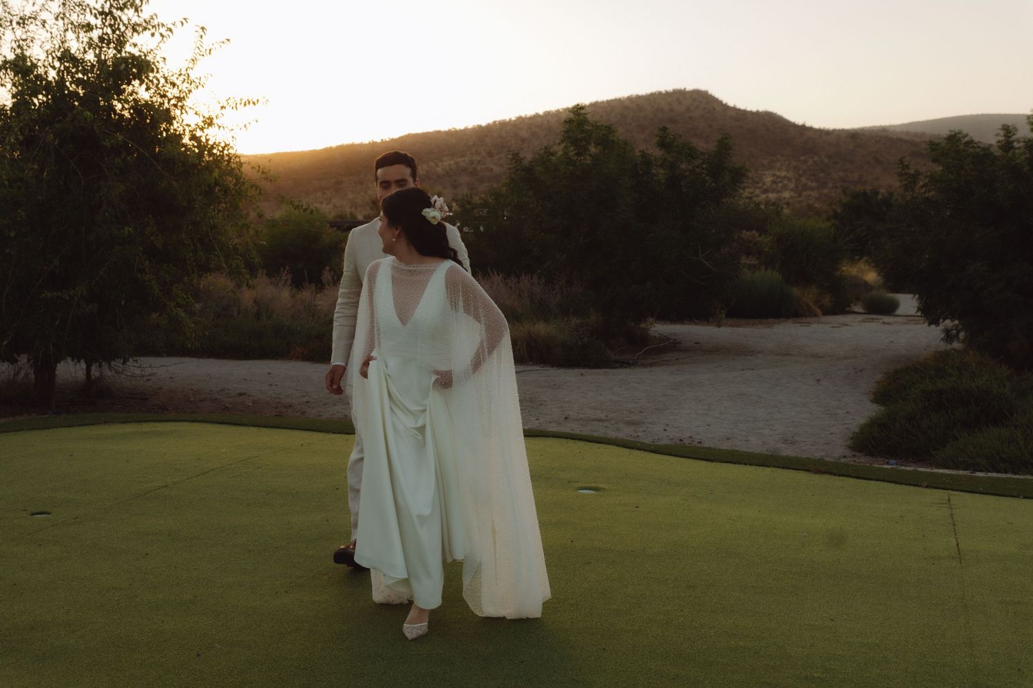 Bride and groom walking hand in hand on lush golf course at golden sunset with mountain backdrop.