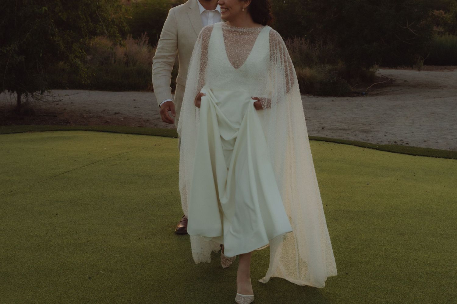 Bride in elegant white gown with flowing cape walking with groom on green golf course at sunset.