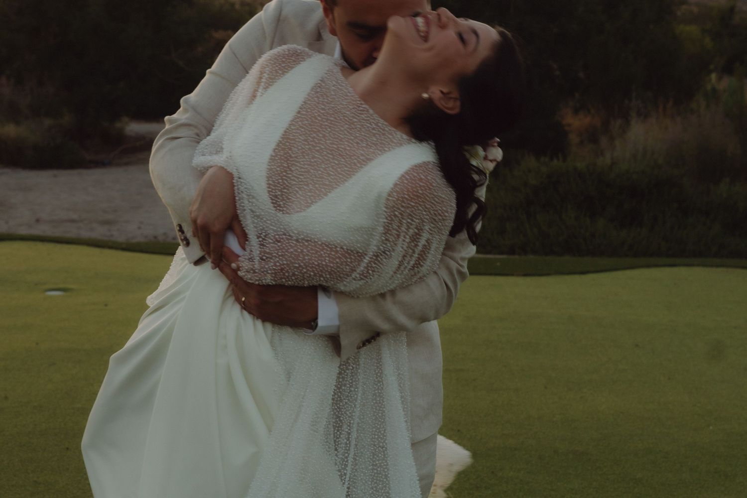 Groom lifting and kissing bride on golf course at dusk, bride in white lace-sleeved wedding gown.