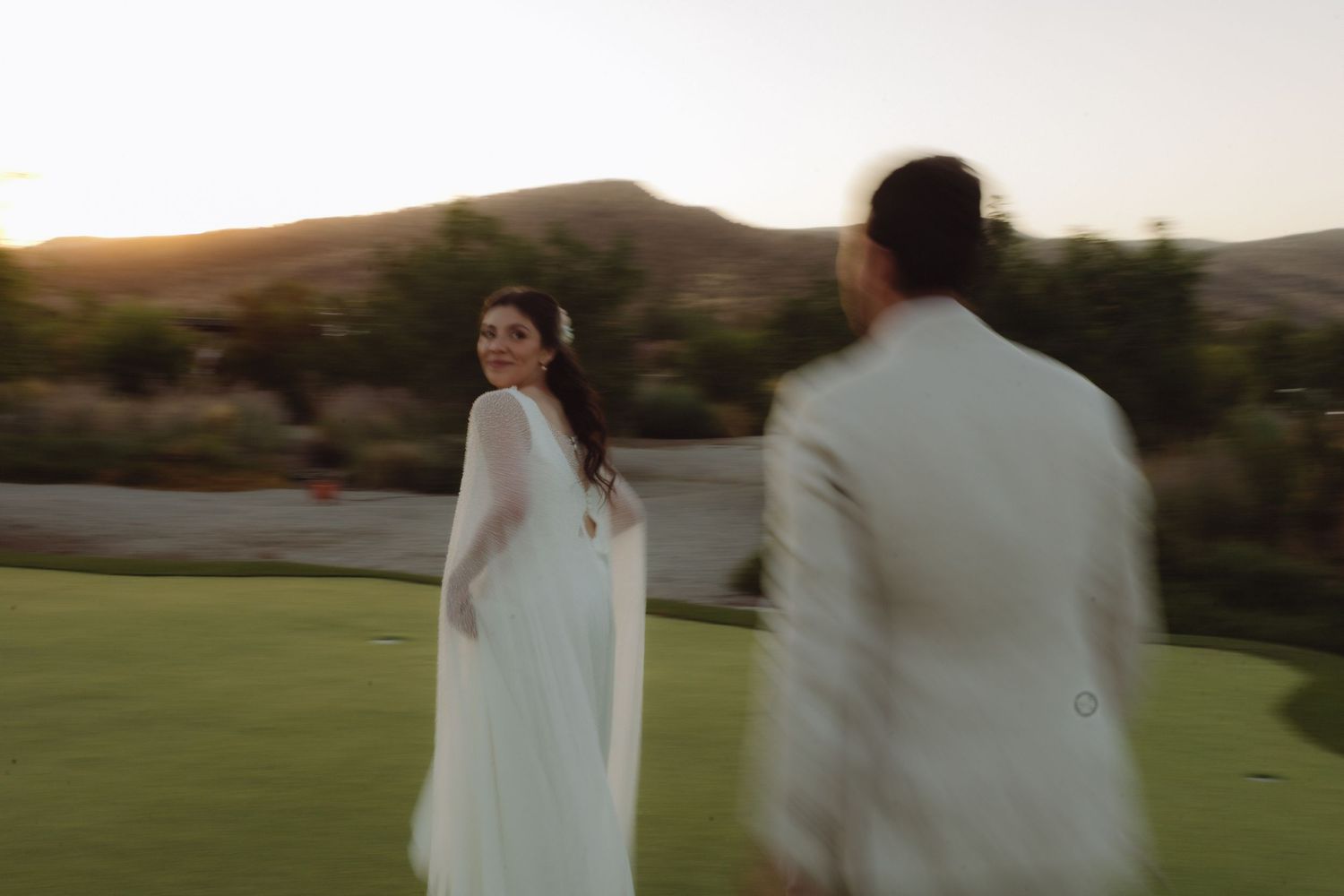Bride in white cape gown glancing back at groom on golf course with warm sunset mountain backdrop.