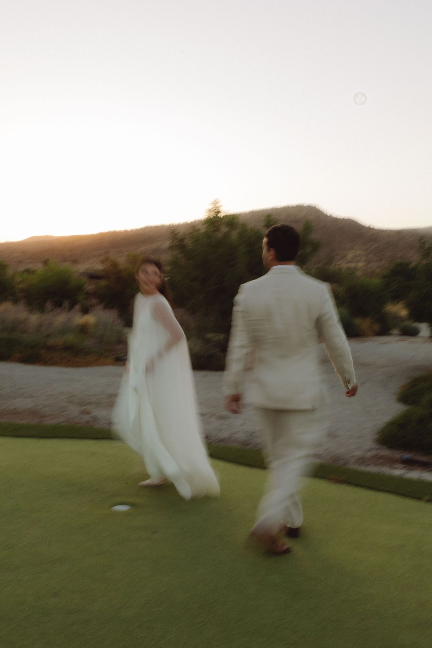 Bride and groom walking hand in hand across green lawn at sunset wedding venue.