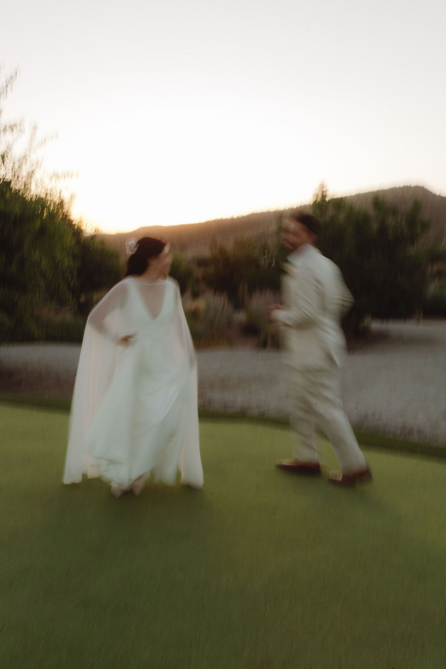 Bride in white cape gown and groom in beige suit strolling on grass at dusk.