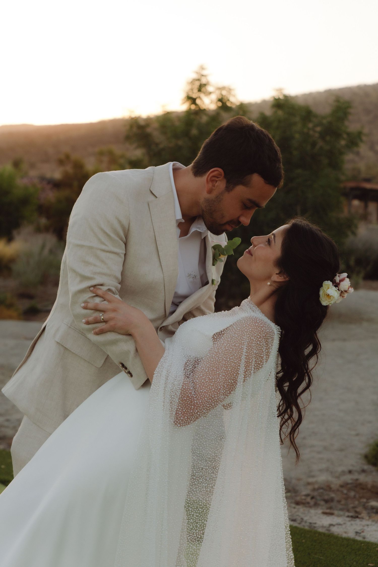 Groom in tan suit dipping bride in pearl cape wedding dress at golden hour.